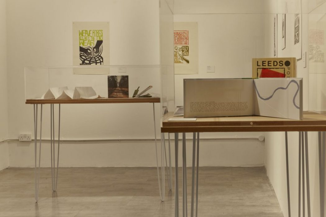An art exhibition features two display tables with books and paper models enclosed in glass cases. Framed posters hang on the walls behind, including one with the word "LEEDS" in bold letters.