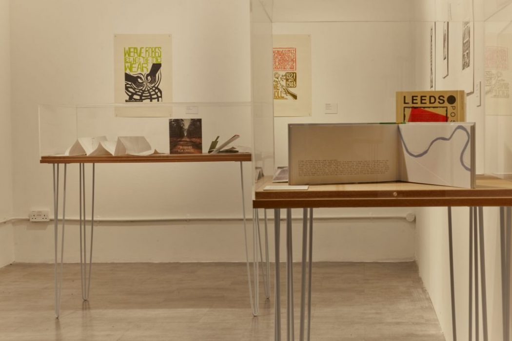 An art exhibition features two display tables with books and paper models enclosed in glass cases. Framed posters hang on the walls behind, including one with the word "LEEDS" in bold letters.