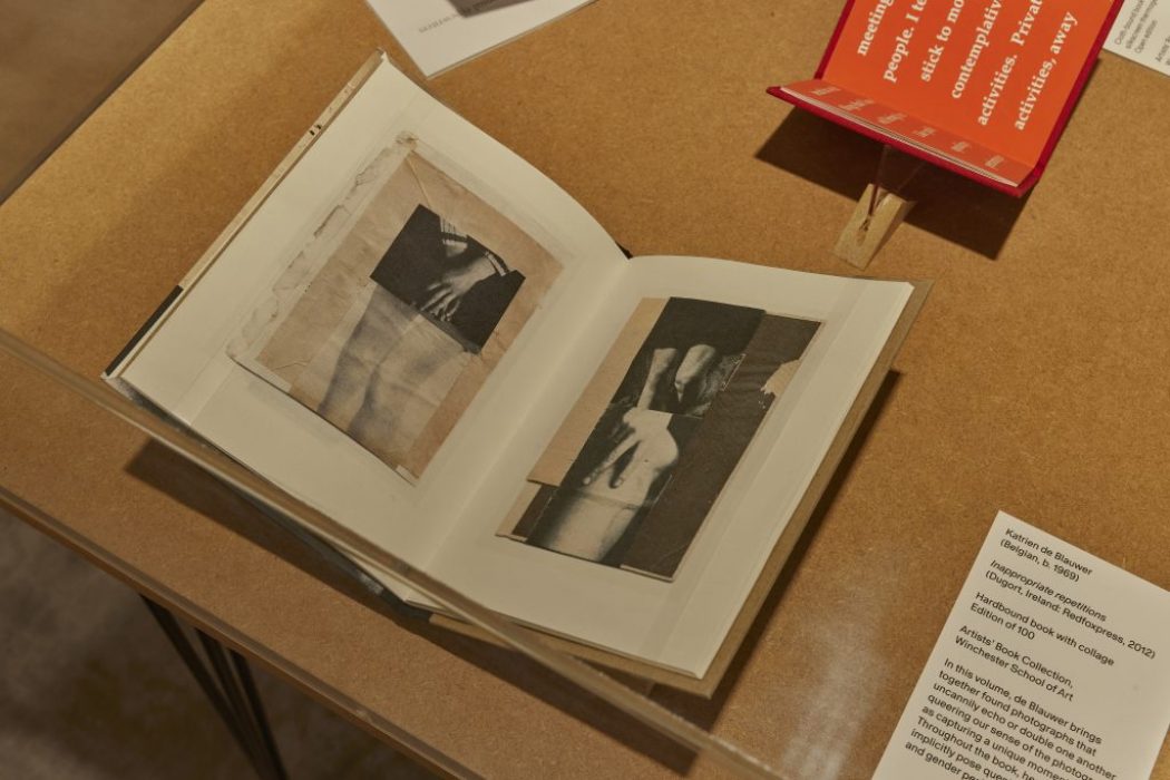 An open book on a wooden table shows black and white images of hands and arms across two pages. A red booklet and a white card with exhibit information are placed beside it.