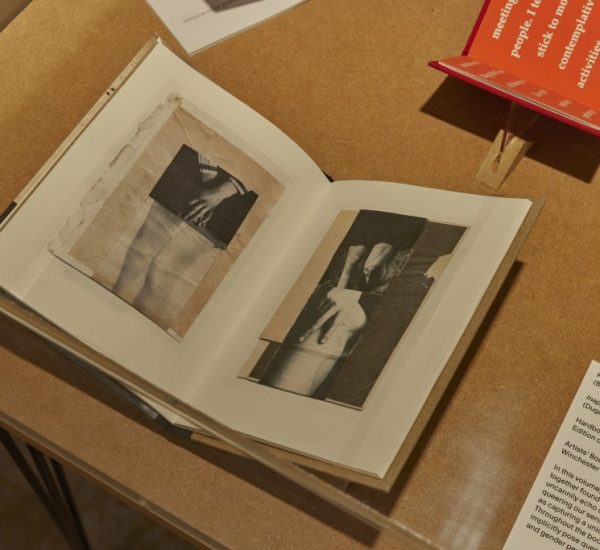 An open book on a wooden table shows black and white images of hands and arms across two pages. A red booklet and a white card with exhibit information are placed beside it.