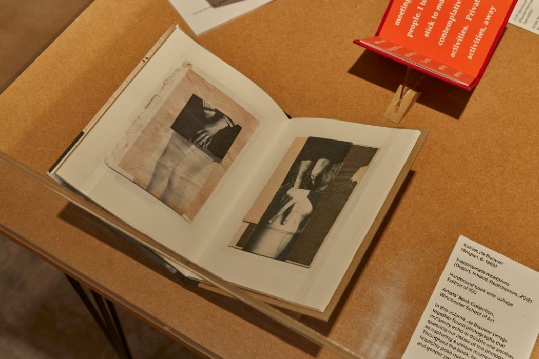 An open book on a wooden table shows black and white images of hands and arms across two pages. A red booklet and a white card with exhibit information are placed beside it.