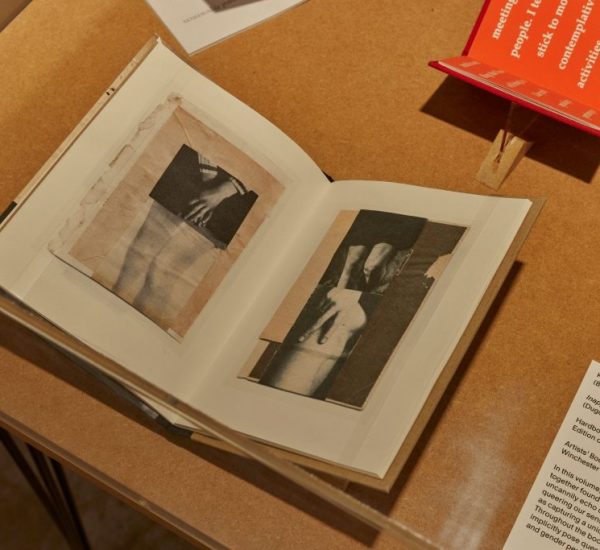An open book on a wooden table shows black and white images of hands and arms across two pages. A red booklet and a white card with exhibit information are placed beside it.