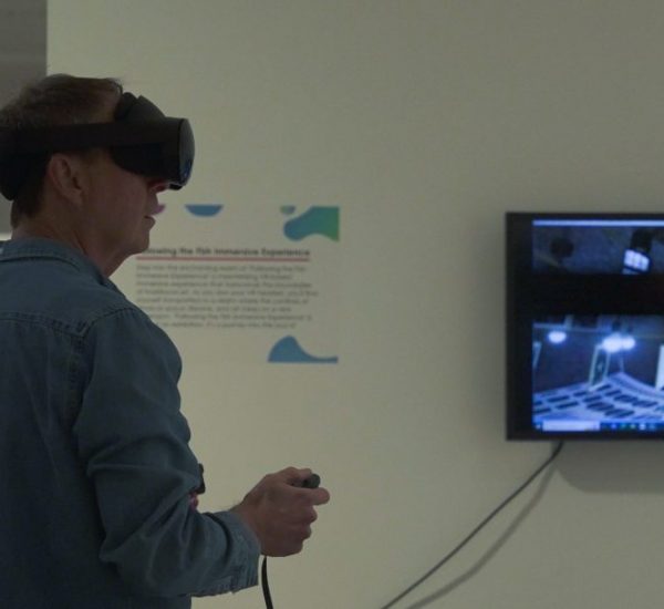 A person wearing a virtual reality headset and holding controllers interacts with a digital experience in an exhibition space. A large monitor on the wall displays a grayscale virtual environment with architectural elements.