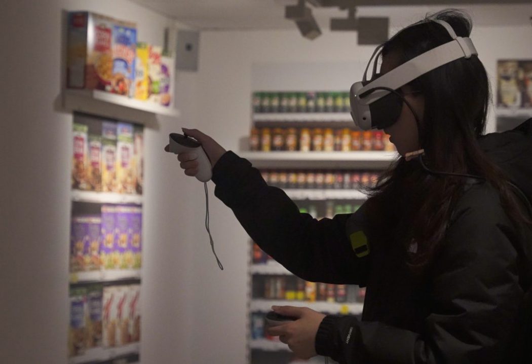 A person wearing a virtual reality headset and holding controllers interacts with a simulated environment in a room designed to look like a grocery store aisle. Shelves in the background are stocked with colorful cereal boxes and jarred goods.
