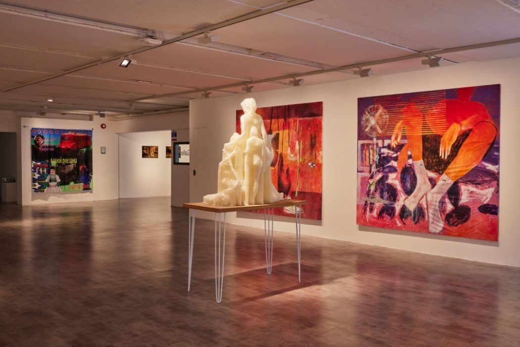 A gallery space shows a large cream-colored sculpture on a table, surrounded by vibrant abstract paintings in red and purple tones.