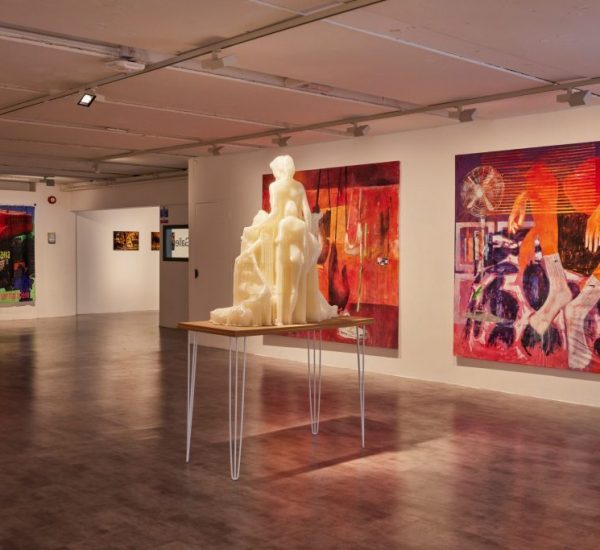 A gallery space shows a large cream-colored sculpture on a table, surrounded by vibrant abstract paintings in red and purple tones.