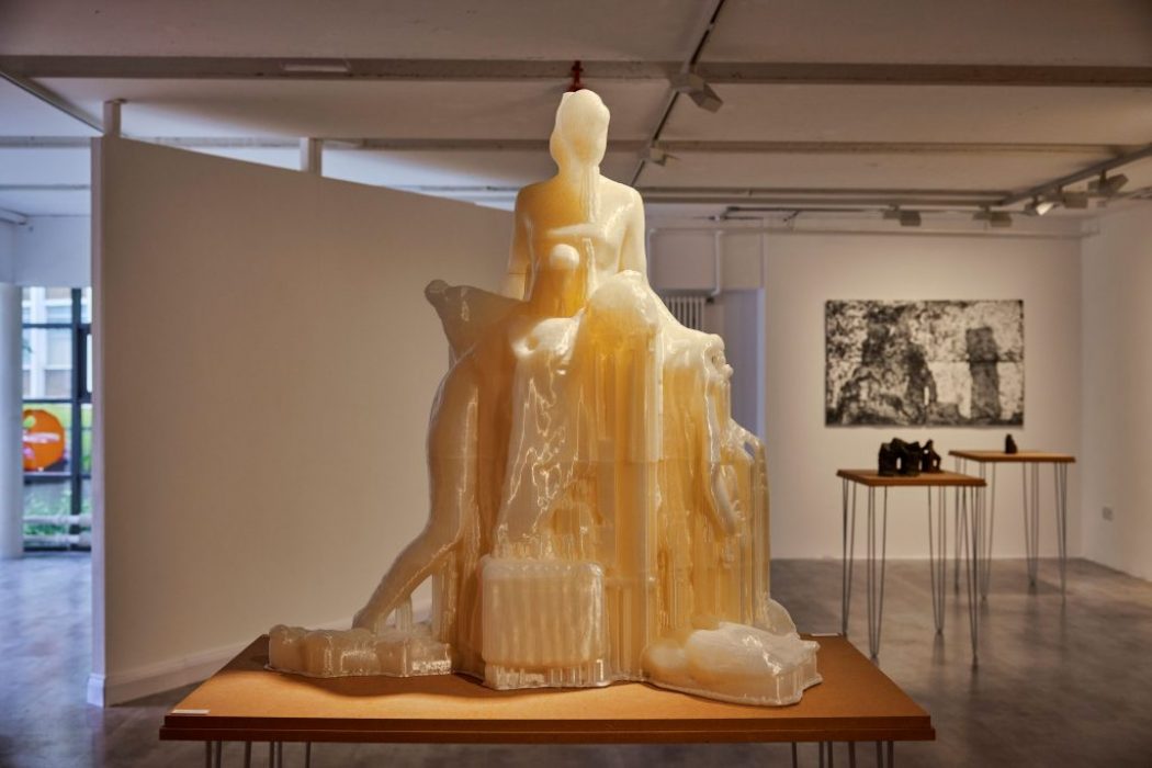 A translucent sculpture of a seated figure with abstract forms is displayed on a wooden table in a gallery, with monochrome artwork and objects in the background.