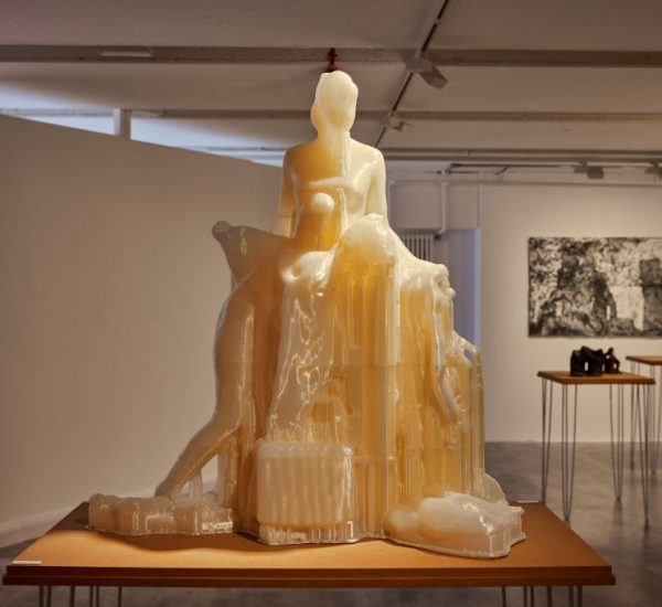 A translucent sculpture of a seated figure with abstract forms is displayed on a wooden table in a gallery, with monochrome artwork and objects in the background.