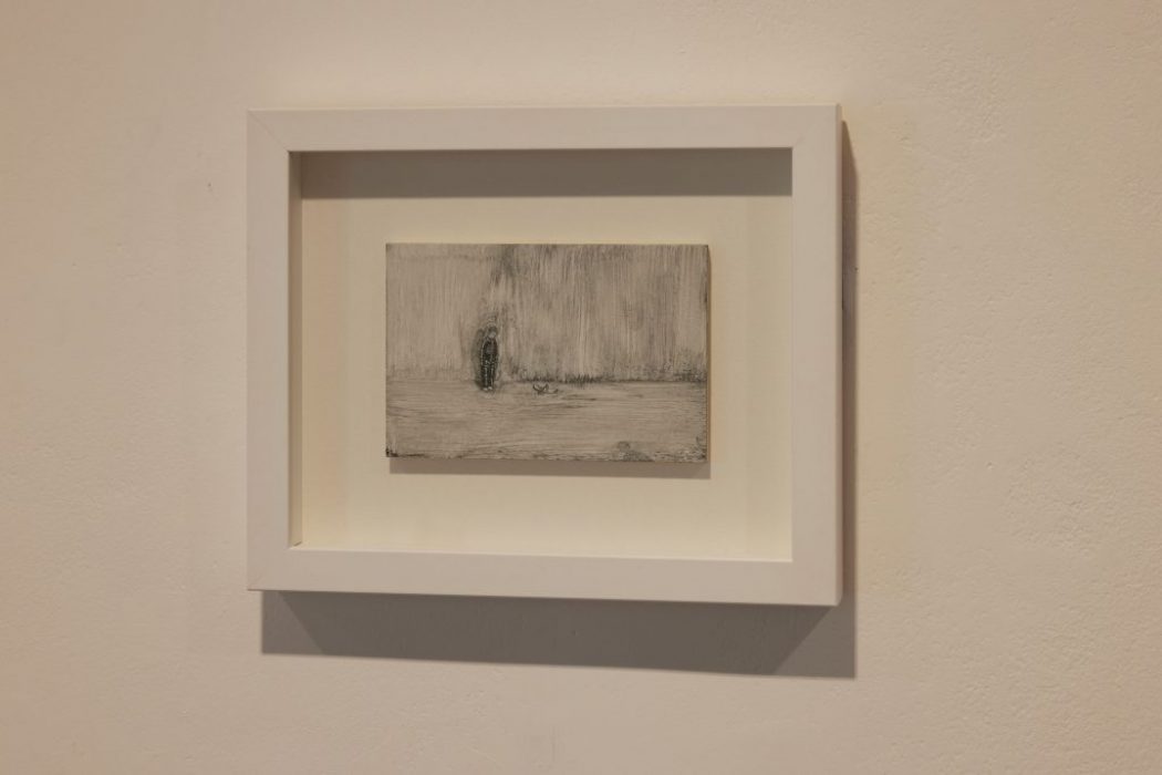 A small monochrome artwork in a white frame depicts a textured scene with a solitary figure, mounted on a plain wall.