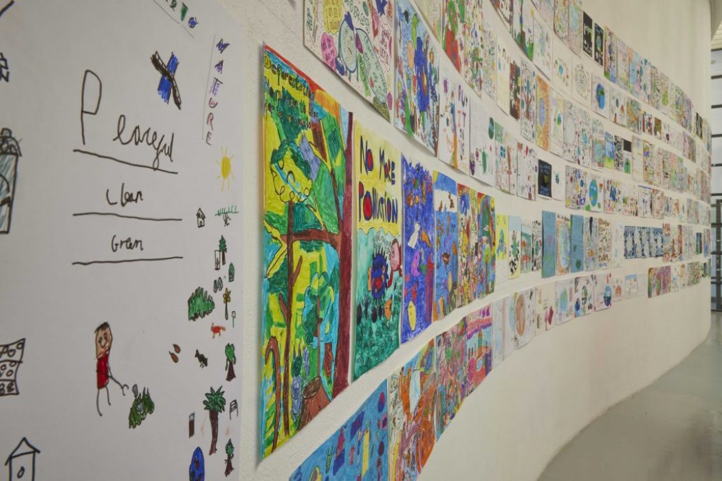 A close-up view of a curved wall shows vibrant children’s drawings, including messages such as “No More Pollution” and nature scenes. The display emphasizes creativity and environmental awareness.