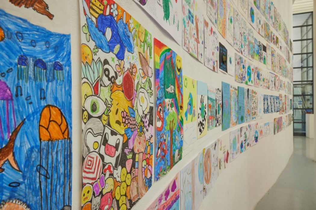 Another close-up of the wall highlights bright, detailed illustrations of animals, plants, and ocean life among other imaginative designs. The artworks are arranged in rows along the curved gallery space.
