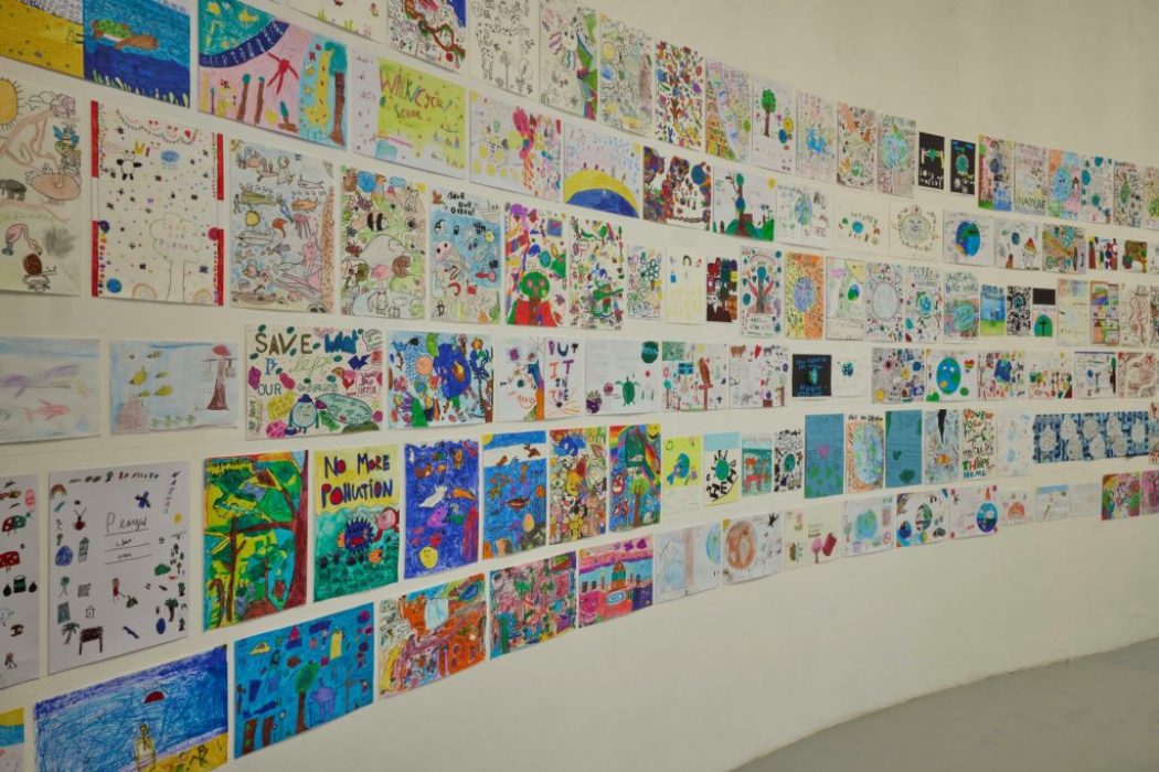 A curved gallery wall is covered with colorful drawings and posters created by children, featuring themes like nature and environmental messages. The artworks are arranged in neat rows across the white surface.