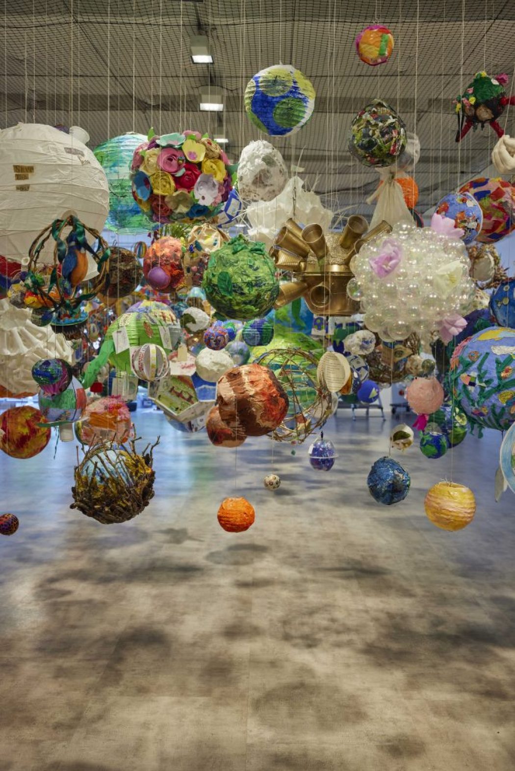 An art installation features numerous colorful spherical objects suspended from the ceiling by thin strings. The spheres vary in size and design, creating a vibrant floating display in a spacious gallery.