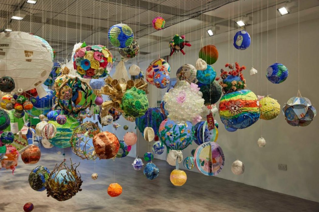 The image shows an art installation featuring numerous colorful spherical objects suspended from the ceiling by thin strings. The spheres vary in size and design, creating a vibrant, floating display in a spacious gallery setting.