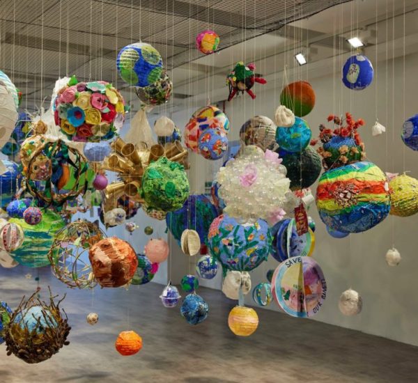 The image shows an art installation featuring numerous colorful spherical objects suspended from the ceiling by thin strings. The spheres vary in size and design, creating a vibrant, floating display in a spacious gallery setting.