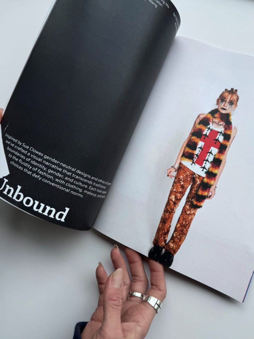 The image shows an open magazine with text on the left page and a fashion photograph on the right. The photograph features a person wearing a sleeveless top with a bold red cross design, a furry striped vest, and patterned trousers.