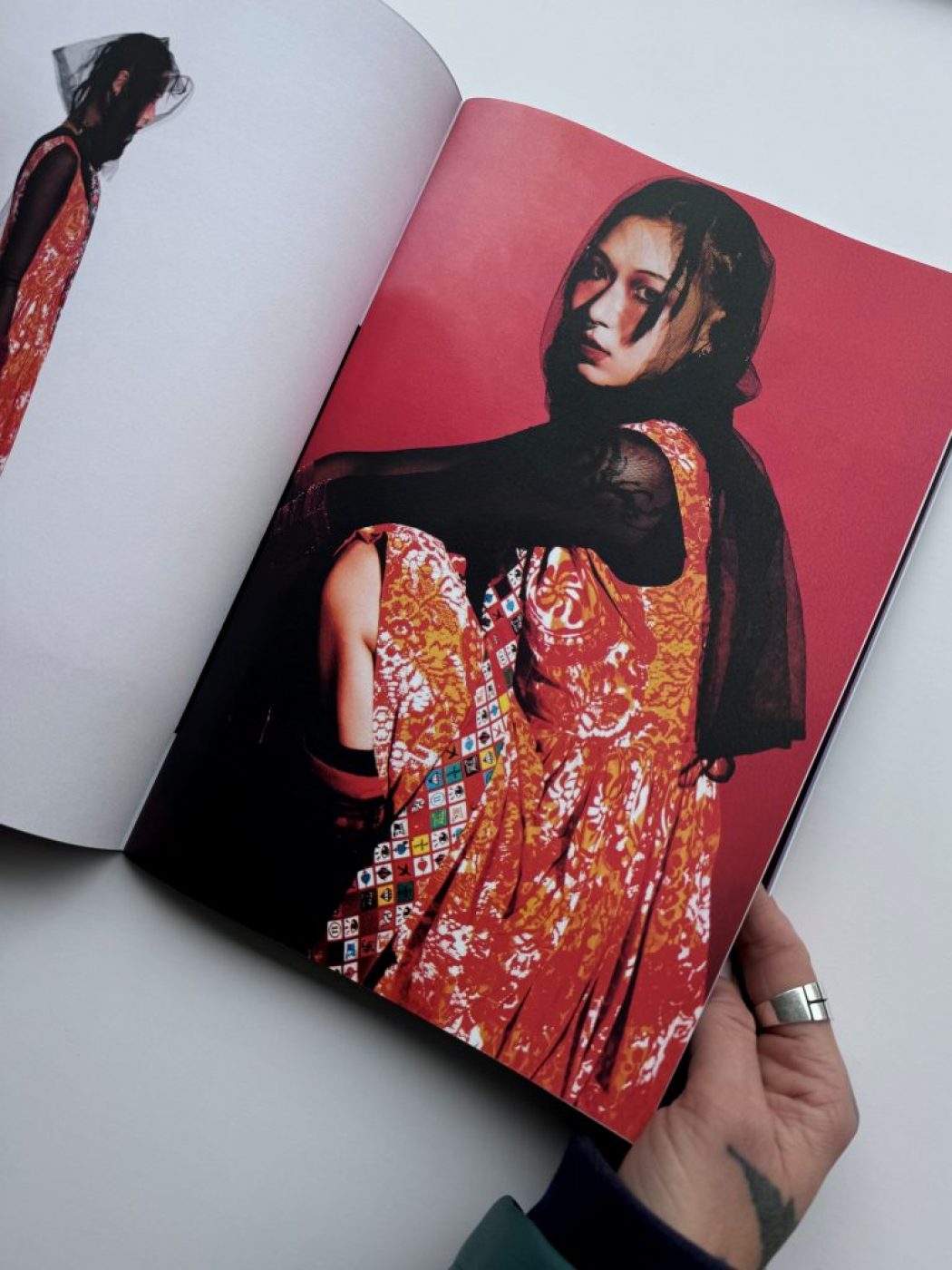 The image shows an open magazine with two fashion photographs against a red background. The outfit features a sleeveless orange patterned dress layered over a black sheer top, accessorized with a colorful geometric sash.
