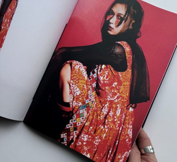 The image shows an open magazine with two fashion photographs against a red background. The outfit features a sleeveless orange patterned dress layered over a black sheer top, accessorized with a colorful geometric sash.