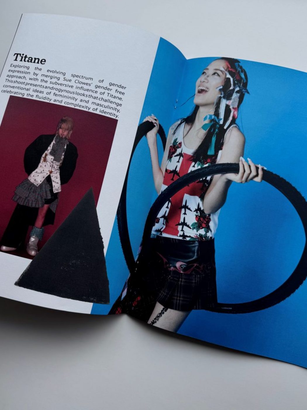 The image shows an open magazine with text on the left page titled “Titane” and a fashion photograph on the right. The photograph features a person wearing a sleeveless patterned top, a short skirt, and holding two large black hoops against a bright blue background.