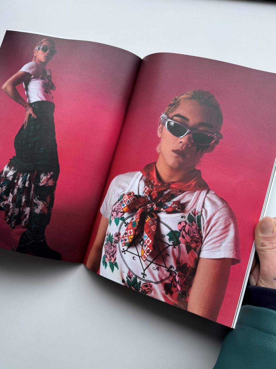 The image shows an open magazine featuring two fashion photographs against a pink background. One page displays a full-body shot of a person wearing a white top, patterned scarf, and a long floral skirt, while the other page shows a close-up of the same outfit.