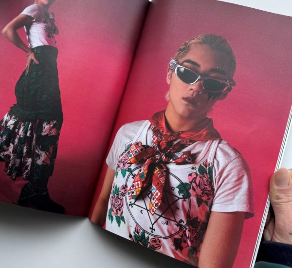 The image shows an open magazine featuring two fashion photographs against a pink background. One page displays a full-body shot of a person wearing a white top, patterned scarf, and a long floral skirt, while the other page shows a close-up of the same outfit.
