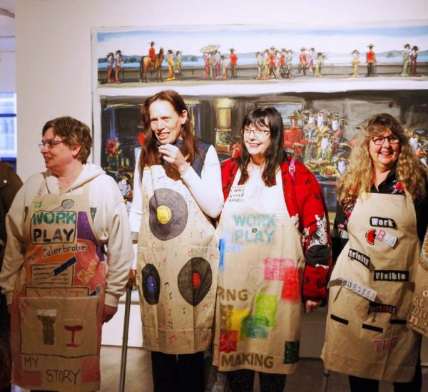 Six people stand in a gallery wearing aprons decorated with colorful text and images, each featuring unique designs and words like “WORK PLAY,” “MY STORY,” and “Adventure.” Behind them, a large horizontal painting depicts a busy outdoor scene with figures and vibrant colors.