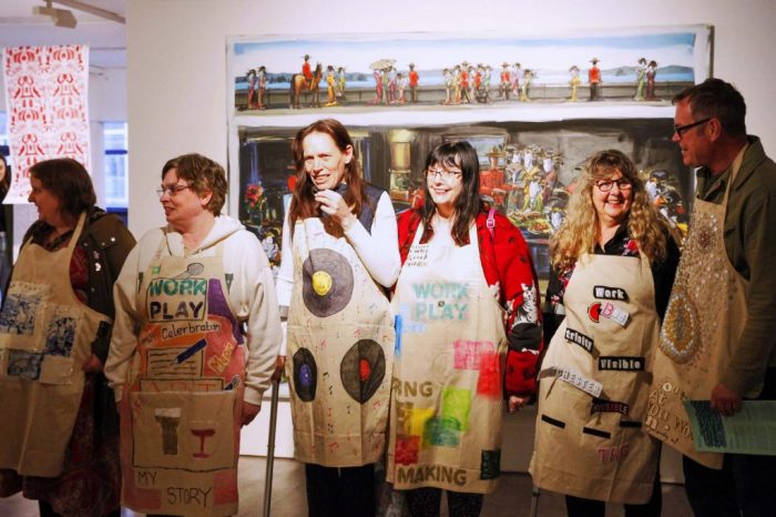 Six people stand in a gallery wearing aprons decorated with colorful text and images, each featuring unique designs and words like âWORK PLAY,â âMY STORY,â and âAdventure.â Behind them, a large horizontal painting depicts a busy outdoor scene with figures and vibrant colors.