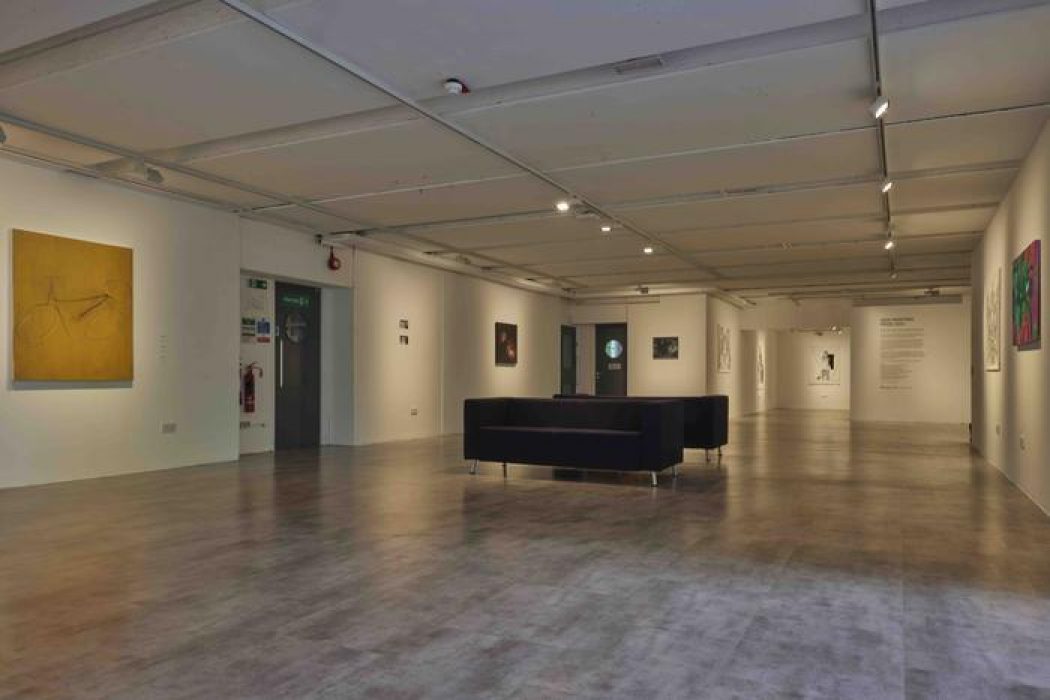 A wide view of the gallery shows multiple paintings on white walls, including a yellow canvas with a faint bicycle outline and vibrant abstract pieces. Black seating is arranged in the center of the room under track lighting.
