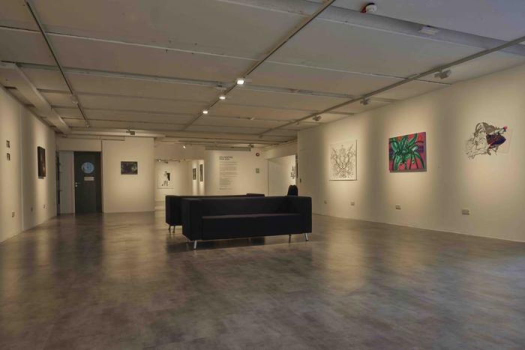 A spacious gallery interior features artworks on both walls, including a green palm-like painting and intricate black line drawings. Two black sofas sit in the center, creating a viewing area under bright ceiling lights.