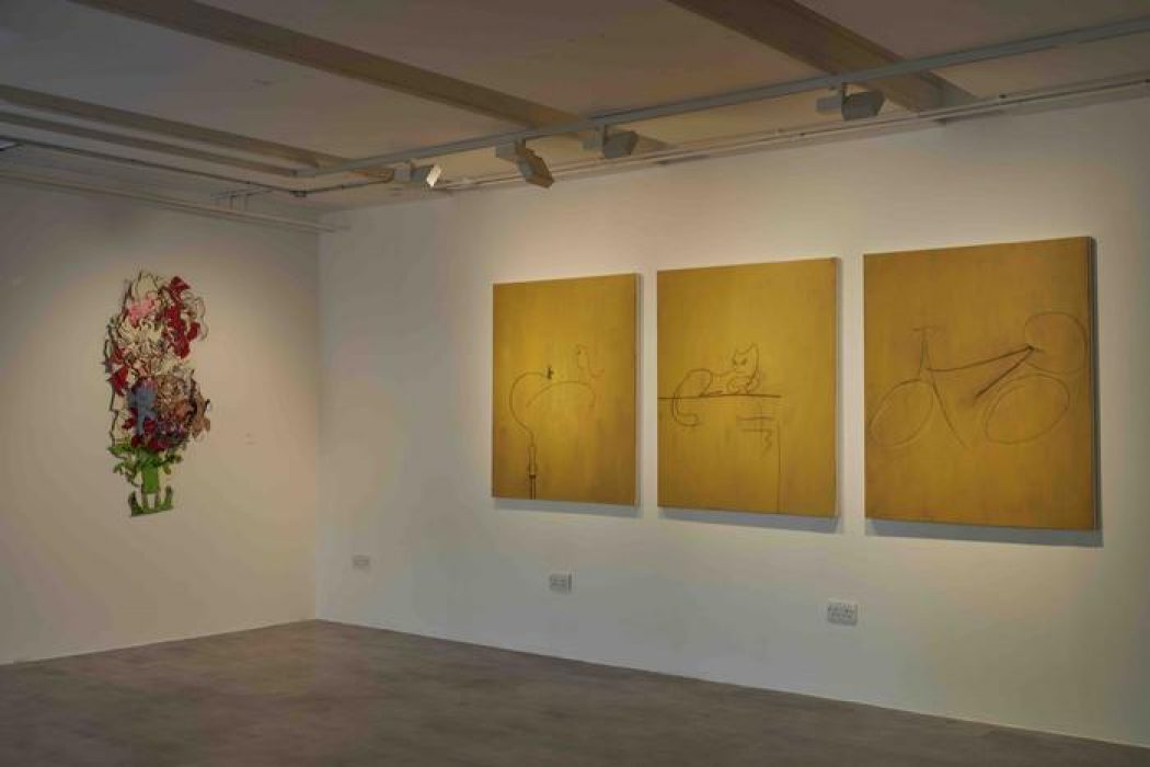 Three yellow-toned paintings with minimalist line drawings of bicycles and animals are mounted on a wall next to a colorful abstract artwork. The gallery floor is gray and the lighting highlights each piece.