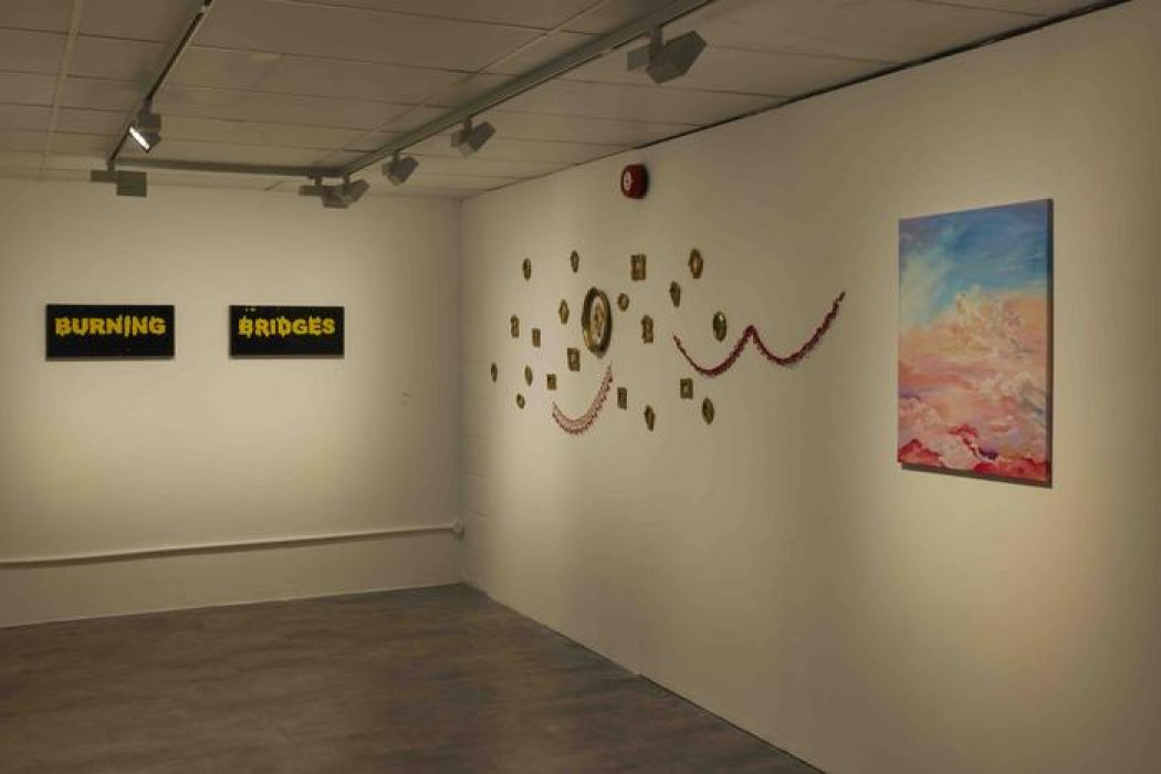 A gallery wall displays two black-and-yellow text artworks reading “Burning Bridges,” a colorful abstract painting of clouds, and a decorative arrangement of small framed pieces with red garlands.