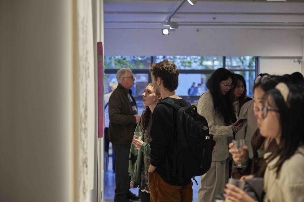 A group of people stand closely together in an art gallery, viewing artworks on the wall and holding drinks.