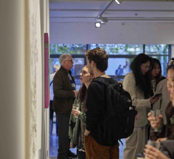 A group of people stand closely together in an art gallery, viewing artworks on the wall and holding drinks.