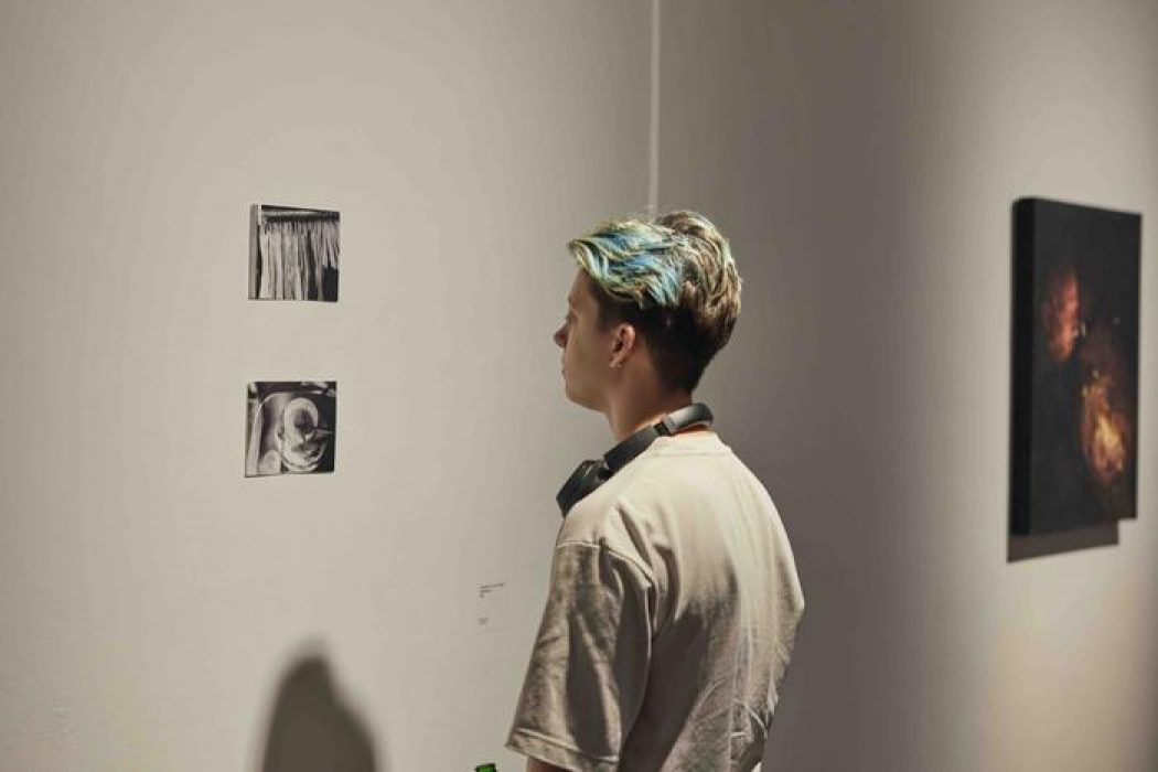 A person observes two small monochrome artworks on a white wall, with a dark abstract painting visible in the background.