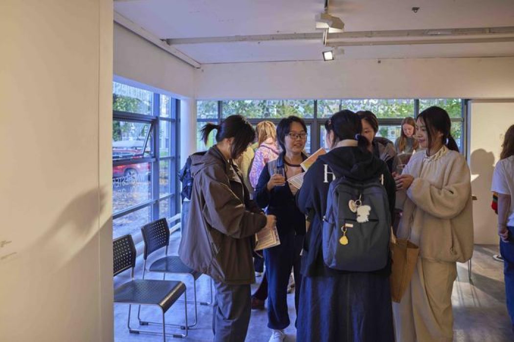 Several people stand near a windowed area of the gallery, holding papers and drinks while conversing.