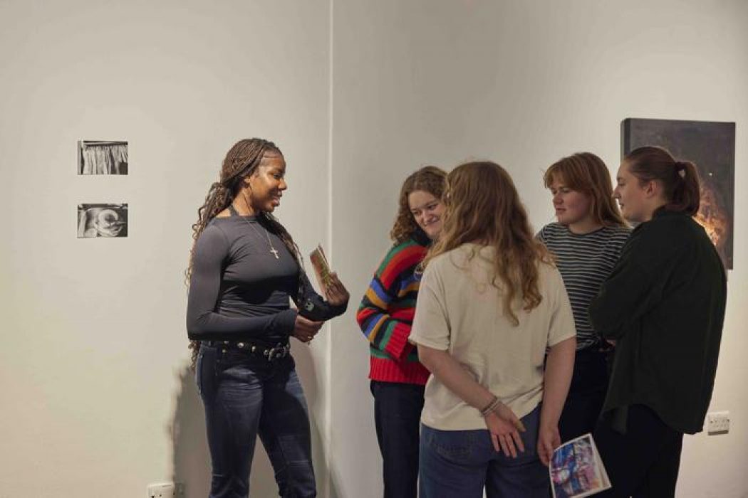A group of people stand in front of a white gallery wall with two small monochrome artworks and a dark abstract painting, holding booklets and discussing.