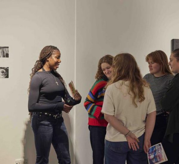 A group of people stand in front of a white gallery wall with two small monochrome artworks and a dark abstract painting, holding booklets and discussing.