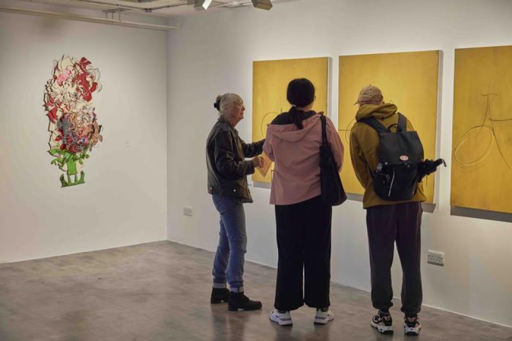 Three people view a series of yellow paintings with simple line drawings on a gallery wall, with a colorful abstract piece visible to the left.