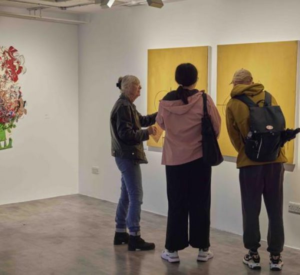 Three people view a series of yellow paintings with simple line drawings on a gallery wall, with a colorful abstract piece visible to the left.