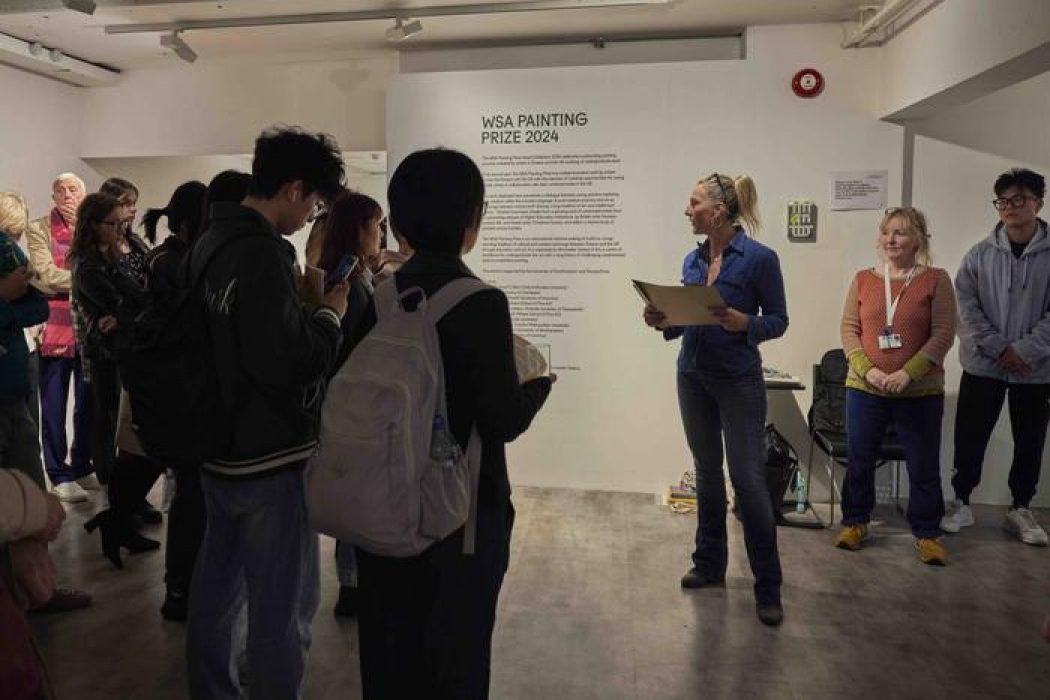 A person speaks to a group in front of a wall with “WSA Painting Prize 2024” text, while others listen and hold papers in the gallery setting.