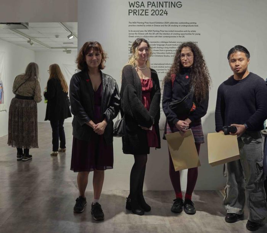Four individuals stand in front of a wall displaying the text “WSA Painting Prize 2024,” holding papers, with other visitors viewing artworks in the background.