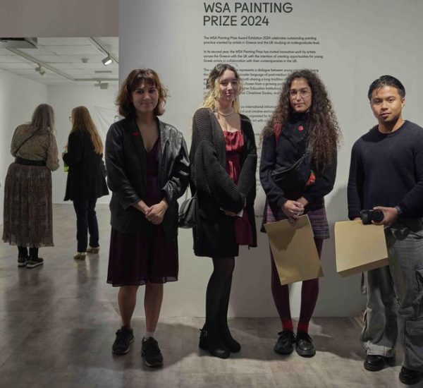 Four individuals stand in front of a wall displaying the text “WSA Painting Prize 2024,” holding papers, with other visitors viewing artworks in the background.