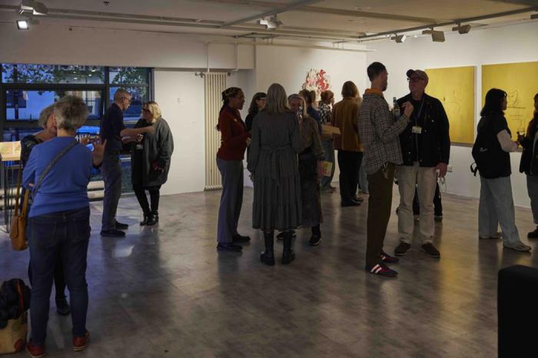 People are gathered in a gallery space, conversing near artworks including two large yellow paintings and a colorful abstract piece on the wall.