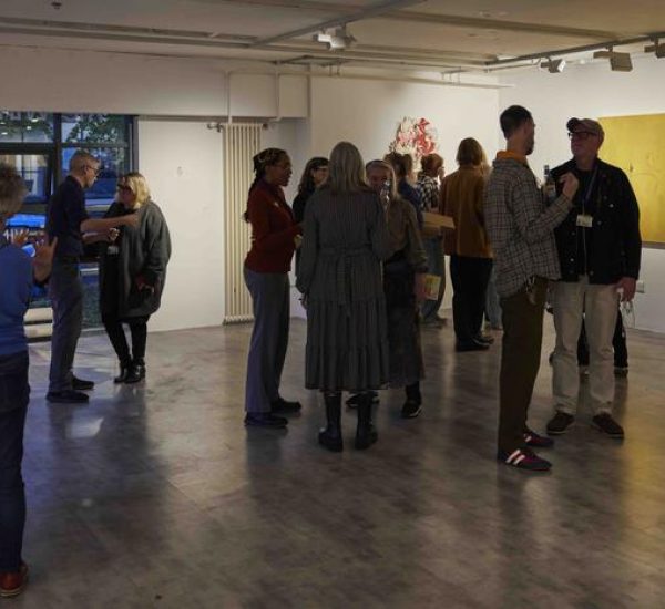 People are gathered in a gallery space, conversing near artworks including two large yellow paintings and a colorful abstract piece on the wall.