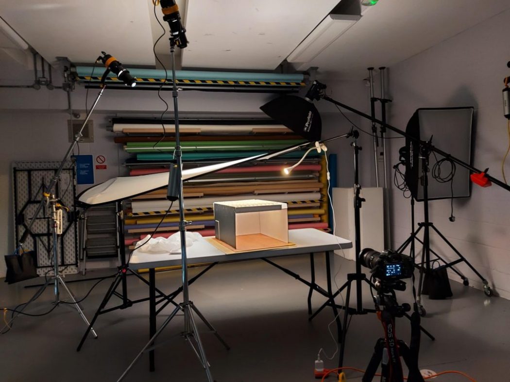 A professional photography studio setup featuring lighting equipment, a camera on a tripod, and a small object on a table being photographed against colorful backdrop rolls.