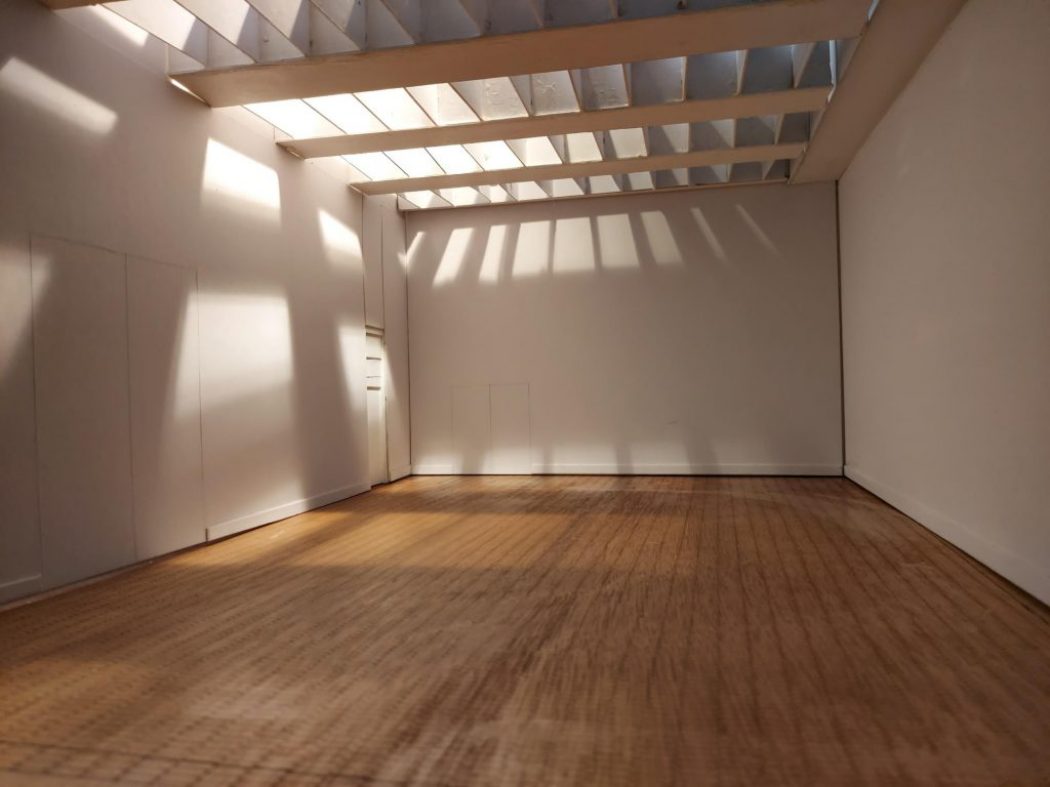 An empty room with white walls, wooden flooring, and a beamed ceiling that casts sunlight patterns across the space.