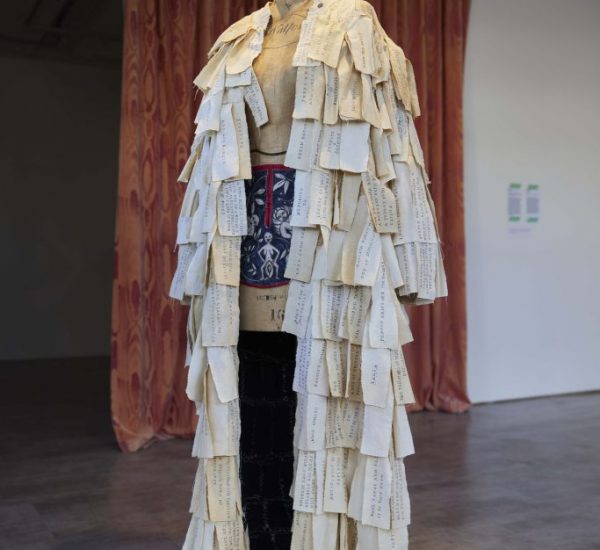 A mannequin stands wearing a long garment made of layered paper pieces with handwritten text. The backdrop includes a patterned curtain, adding texture to the display.