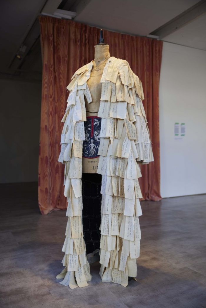 A mannequin stands wearing a long garment made of layered paper pieces with handwritten text. The backdrop includes a patterned curtain, adding texture to the display.