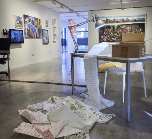 A gallery space features a table with a lamp and long sheets of paper covered in handwritten text spilling onto the floor. Additional artworks hang on the walls in the background, creating a dynamic installation.