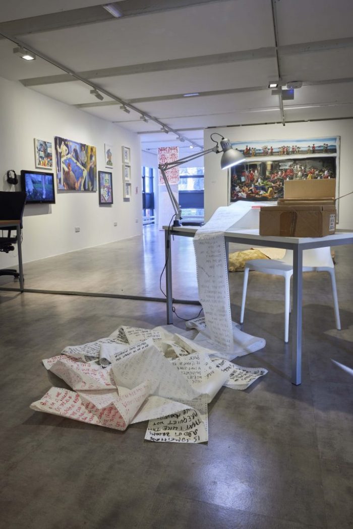 A gallery space features a table with a lamp and long sheets of paper covered in handwritten text spilling onto the floor. Additional artworks hang on the walls in the background, creating a dynamic installation.